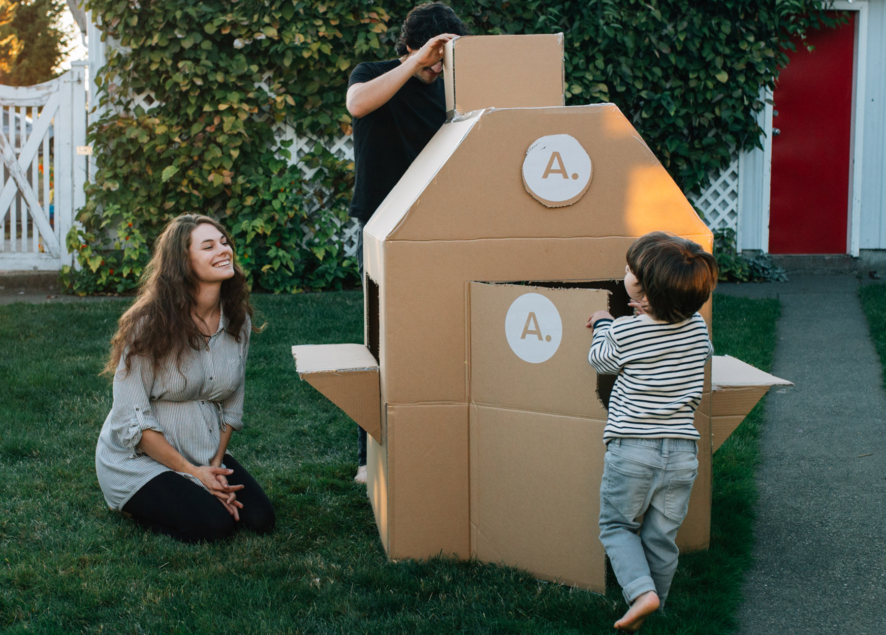 A family enjoys playing with a cardboard box fort. There are awesome details like a chimney, door, and window ledges.